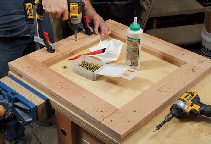 Asa continues to drive screws into a now fully assembled arm. A bottle of glue and a box of screws lay on the bench. There is also a impact driver with a countersink bit visible.